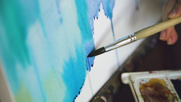 Detailed view of a hand using a paintbrush to apply blue paint on a canvas.